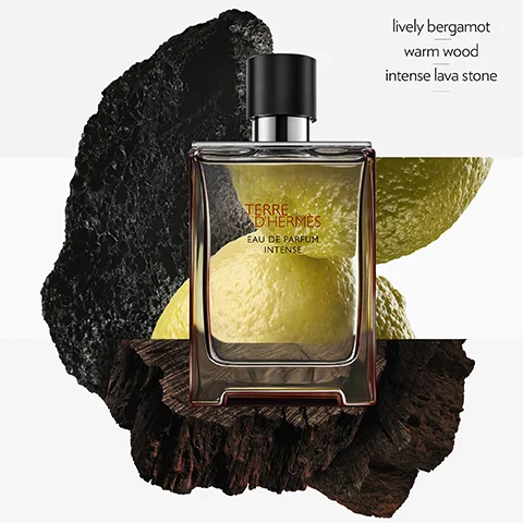 A Terre dHermes Eau de Parfum Intense fragrance bottle with a black cap sits on dark rough wood and lava stone, with blurred yellow-green citrus slices in the background and through the bottle, next to text that reads lively bergamot warm wood intense lava stone.